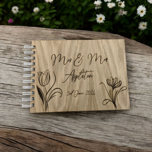 A personalized engraved wooden wedding guest book with floral patterns and names and a date, placed on a wooden surface with a garden in the background.