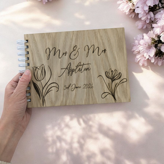 A close-up photograph of a personalized wooden wedding guest book with wire binding, featuring engraved script that reads "Mrs & Mrs Appleton 3rd June 2026" and elegant floral sketches, held by a hand