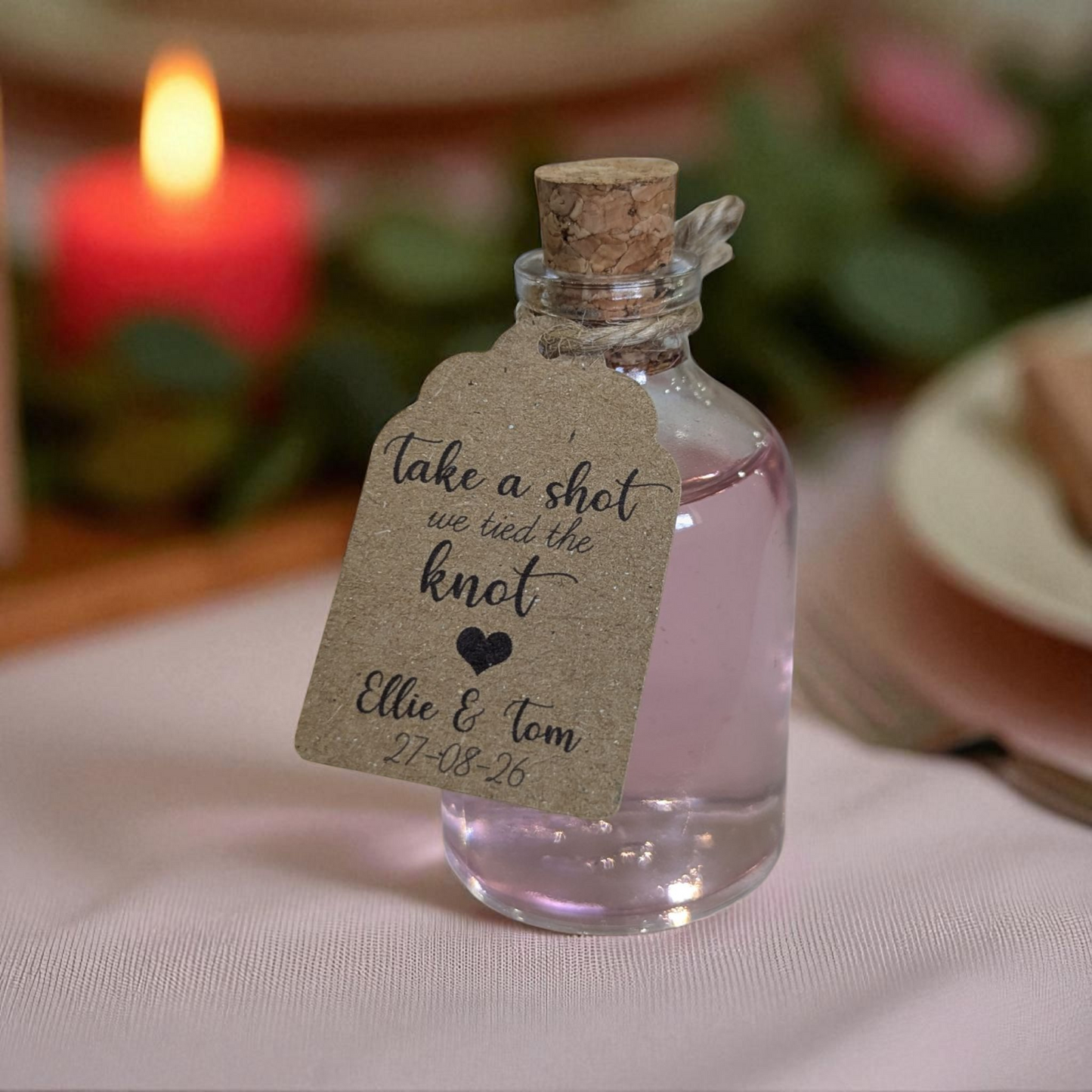Rustic bottle with a cork lid and personalised kraft tag on a wedding table place setting with a blurred candle and plate in the background.