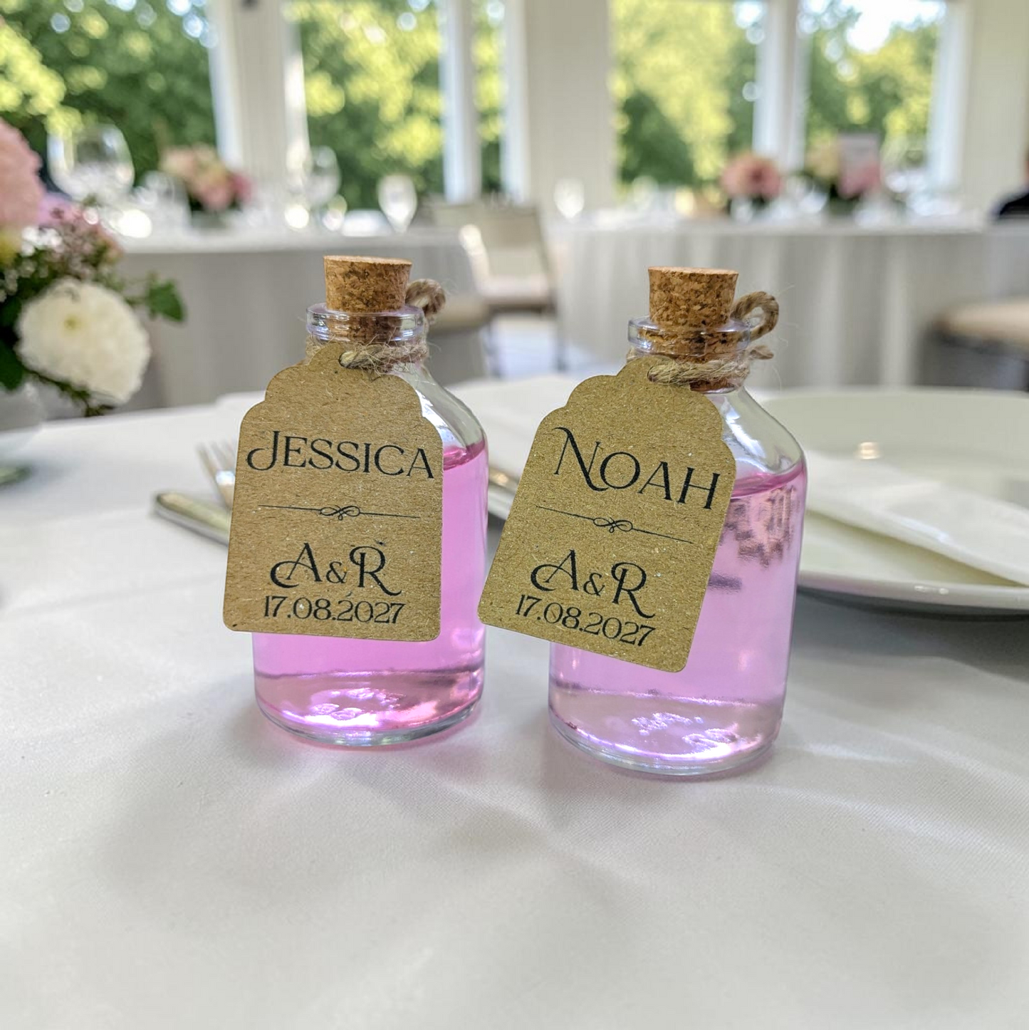 Two small wedding favour bottles with guest's names and couples' initials and wedding date. Intended to be used as a unique wedding favour and table name. Bottles stand in front of white place setting in wedding venue