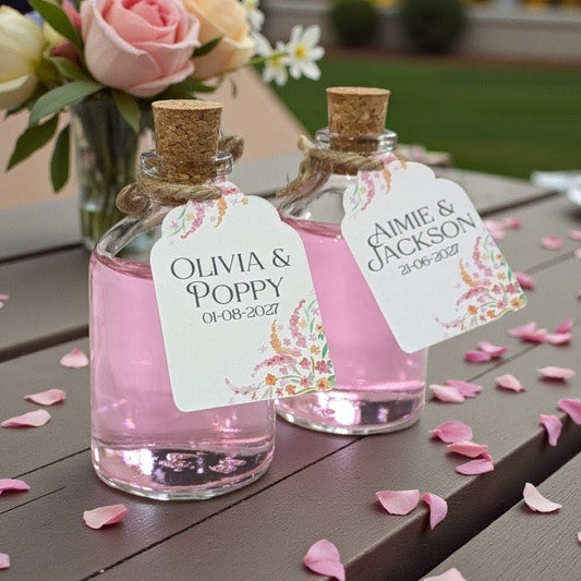 Two small wedding favour bottles with cork stoppers and personalised tags on a wooden surface with pink petals. Being used as custom wedding favours for guests.