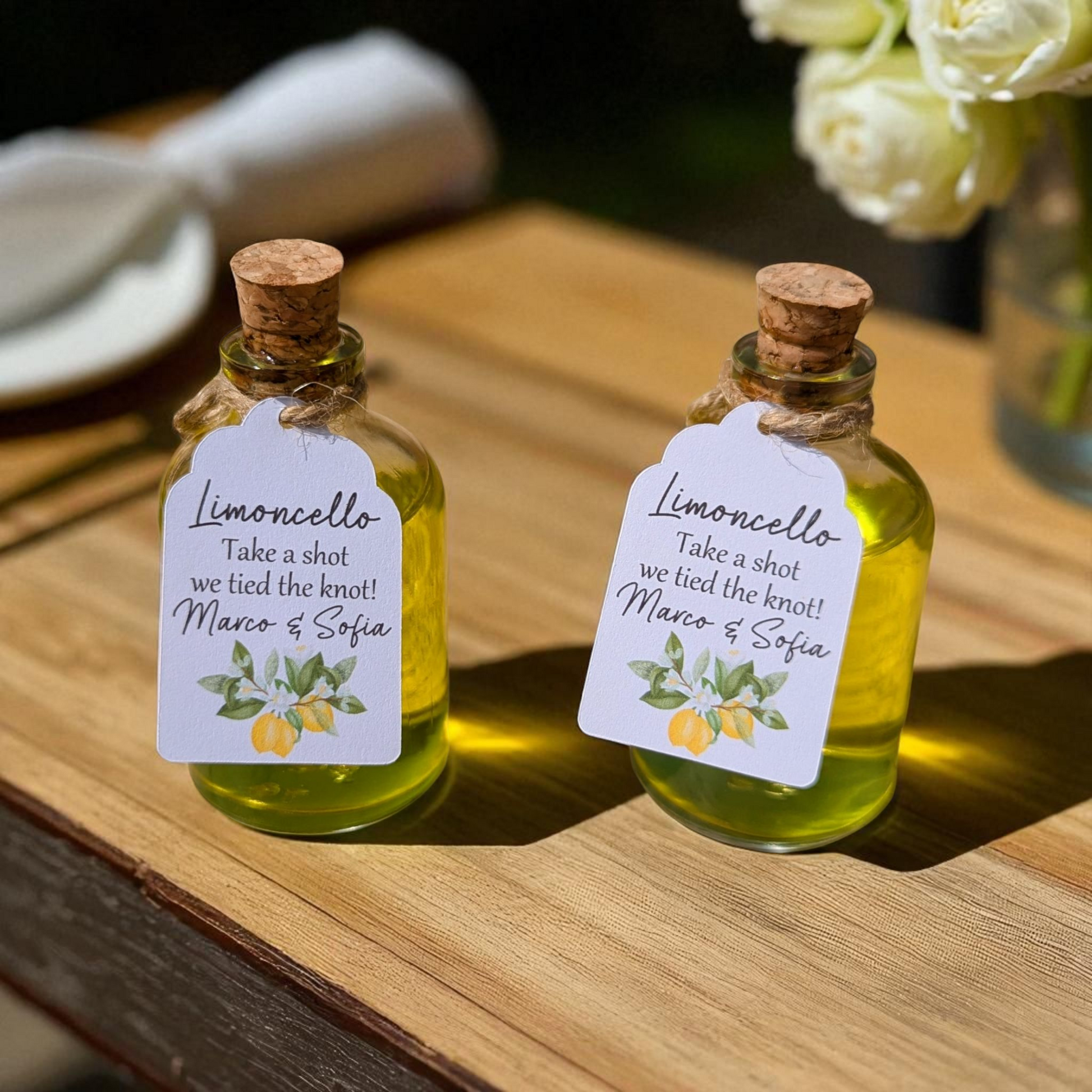bulk wedding favours for guests. Limoncello fillable corked bottles with customisable tags on a wooden surface with wedding decor in the background