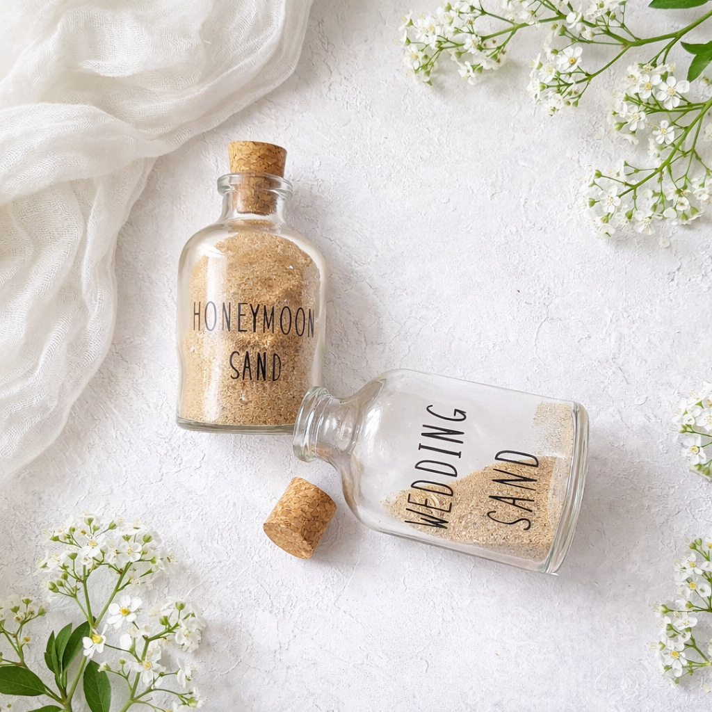 A pair of personalised wedding keepsakes and honeymoon keepsake sand bottles with cork stoppers on a white floral background