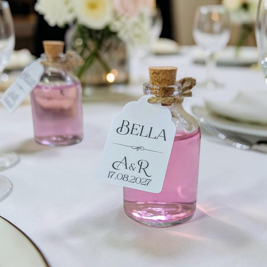 Close-up of a mini glass bottle wedding favour featuring a personalised name card tied with twine on a white wedding table cloth.