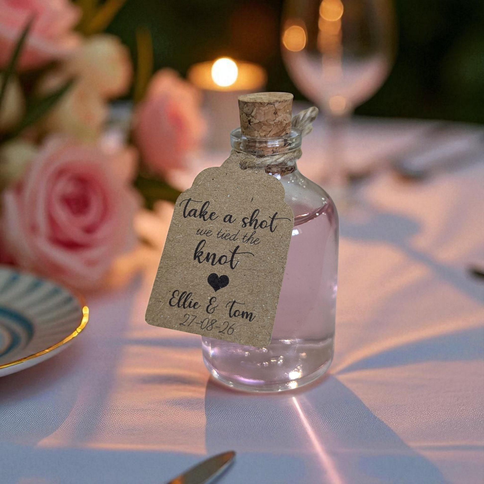 A 50ml glass bottle with a cork top, filled with gin on a wedding table with a personalised tag tied onto the wedding favour. A soft glow comes from a candle behind.