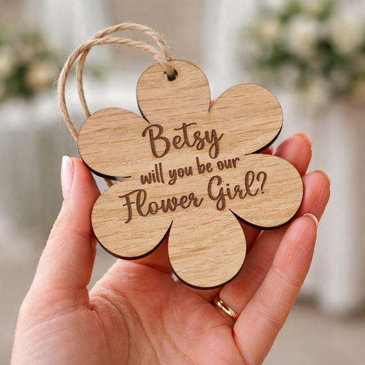 Wooden flower-shaped tag with 'Betsy, will you be our Flower Girl?' text held in a hand.