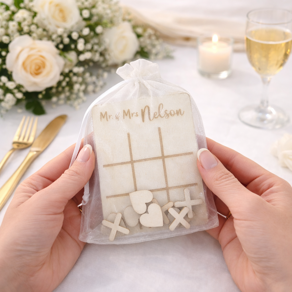 Hand holding an organza wedding favour bag with personalised tic tac toe table game inside, on a table setting with flowers and wine.