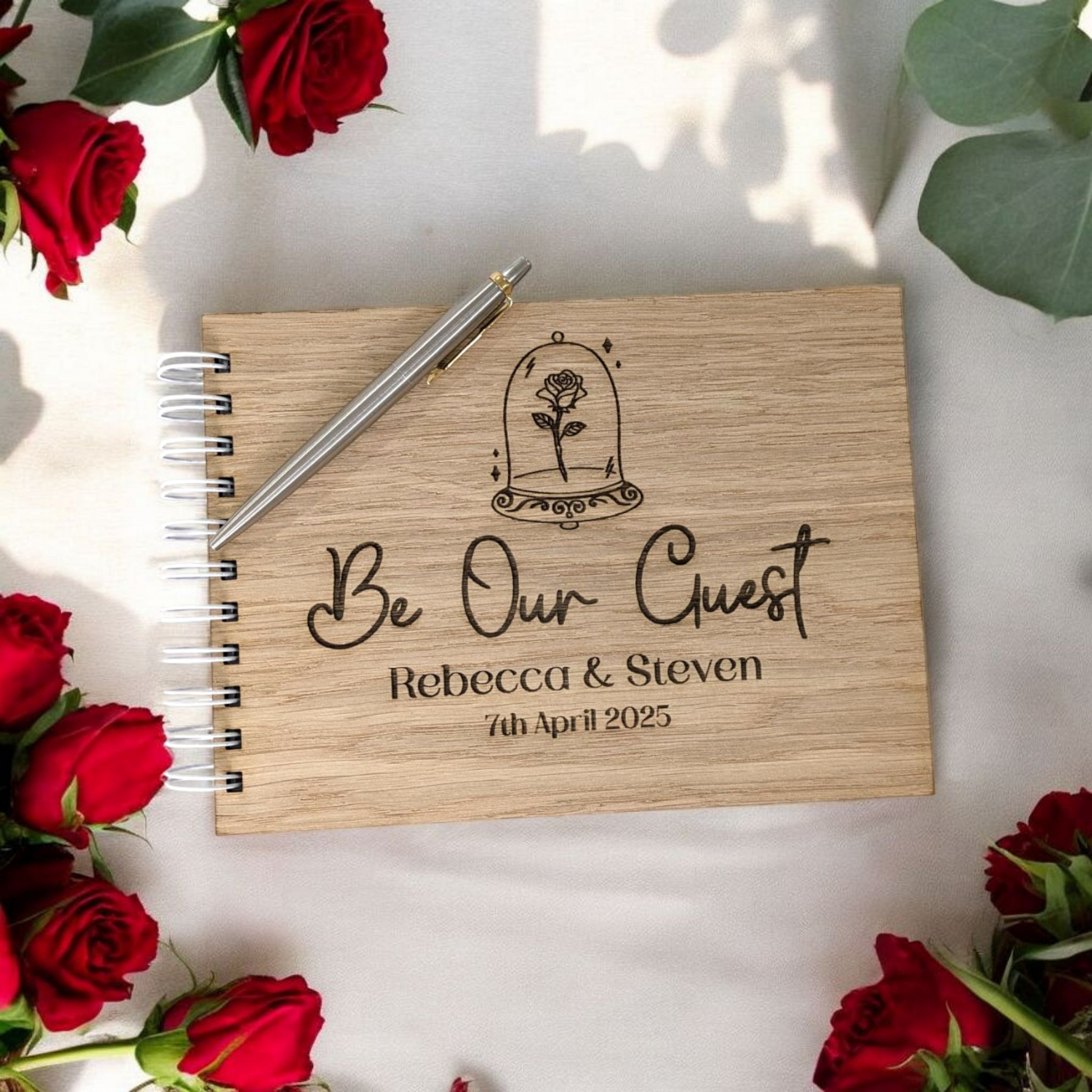 An engraved sign in guest book for a wedding with the phrase 'Be Our Guest' and a rose dome design, personalised with names and a wedding date, displayed on a white surface surrounded by red roses.
