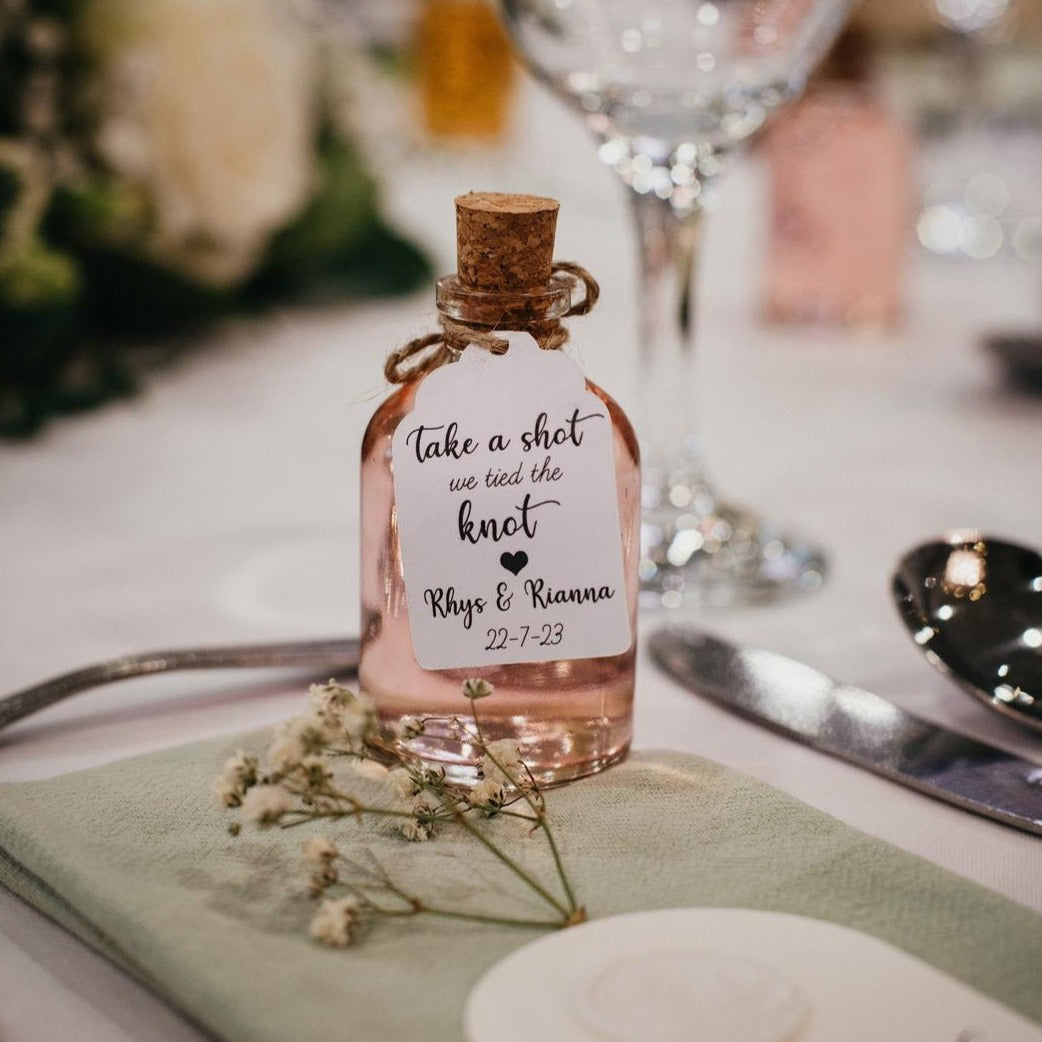 A Wedding Shot Bottle Favour with a personalised tag sat on a sage green napkin at a wedding table setting. The 50ml Glass Bottle is perfect for filling with the bride and groom’s favourite drink.