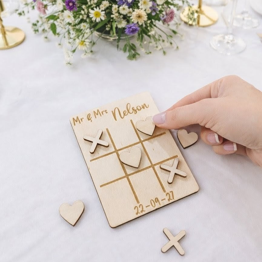 Wooden tic-tac-toe game with personalised 'Mr & Mrs' engraving on a wedding table with white table cloth and flowers in the background.