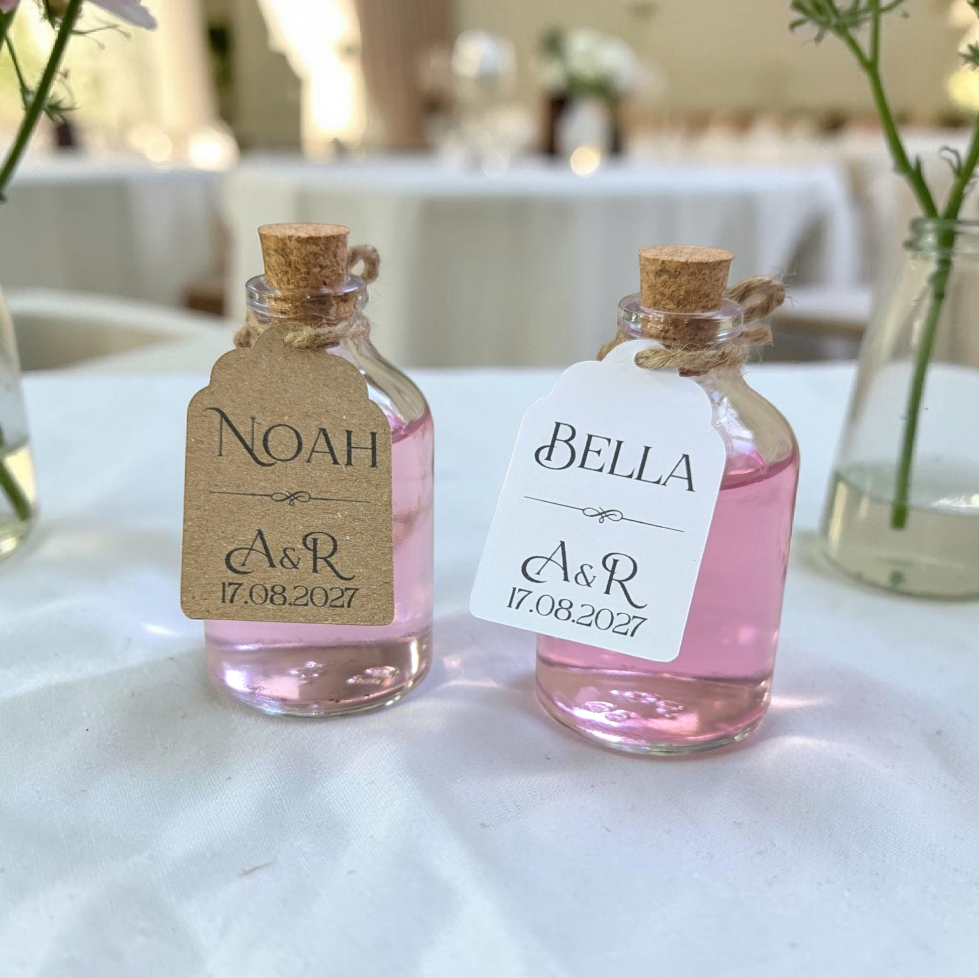 Pair of mini bottles with custom place card tags. Tags are printed with guest name and couple's initials. In the background is a wedding scene.