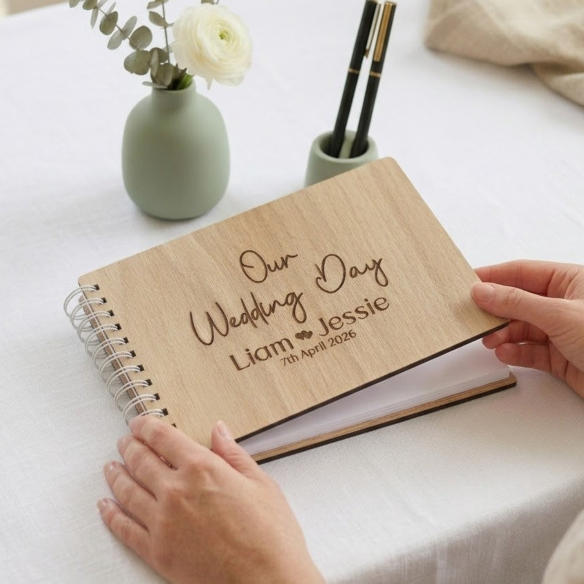 Wooden wedding guest book with engraved text on a white surface with a vase and pens.