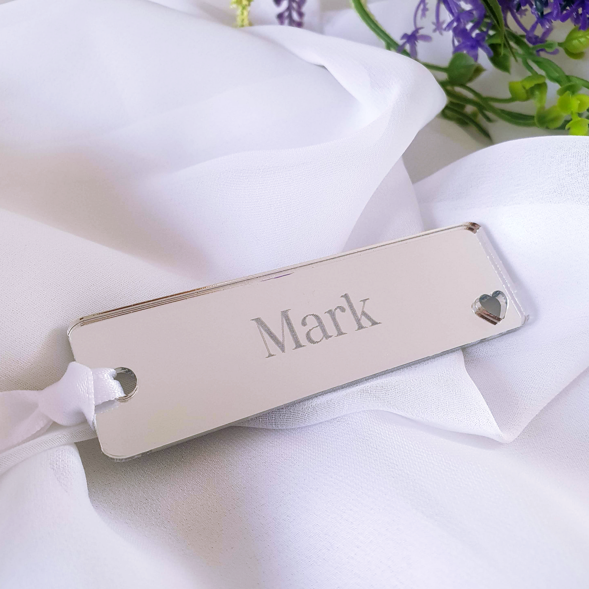 A silver bookmark present with a personalised engraving of a name. Bookmark is custom designed with white ribbon and a heart shaped cut out.